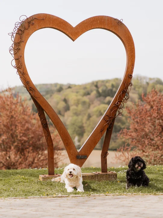 Zwei kleine Hunde liegen vor dem Herz-Aufsteller im Gartenareal vom Hotel das Eisenberg.