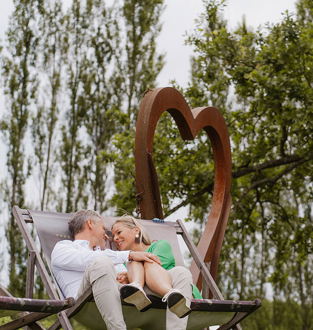Paar sitzt auf einer Holzbank vor dem Herz-Aufsteller im Gartenareal beim Hotel das Eisenberg.